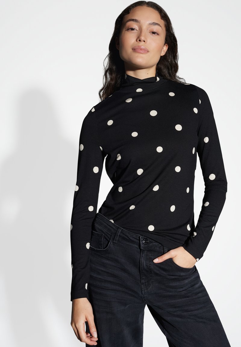 Zwarte lange mouw top met witte stippen, voorzien van een hoge halslijn. Gecombineerd met een donkere denim broek, die een slim fit ontwerp laat zien.