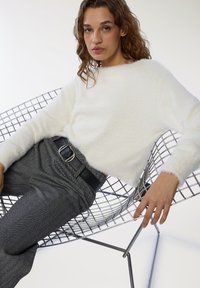 Maglione bianco soffice con scollatura rotonda abbinato a pantaloni grigi a spina di pesce e una cintura nera; seduto su una sedia di filo.