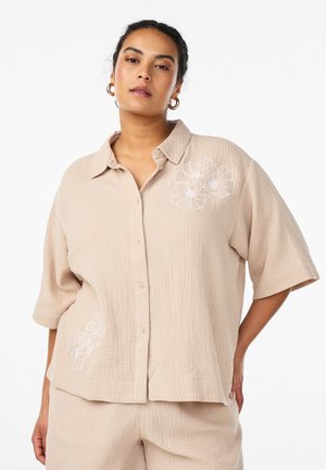 Kobieta w luźnej beżowej koszuli na guziki i spodniach z białym kwiatowym haftem, pozująca na jednolitym białym tle.