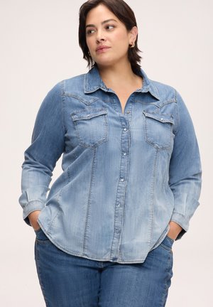 Donna che indossa una camicia di jeans azzurro chiaro con tasche frontali e jeans blu, in piedi con le mani nelle tasche, guardando leggermente di lato.