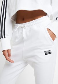 Felpa bianca corta con tre strisce nere sulle maniche, abbinata a pantaloni della tuta bianchi con coulisse e toppa con logo Adidas nero.