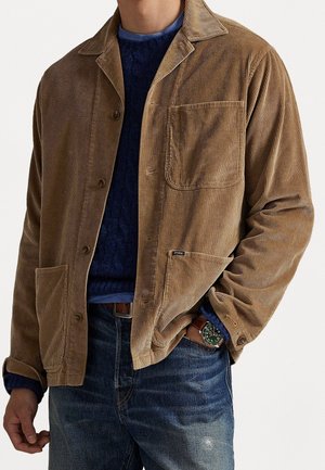 Homme portant une veste en velours marron sur un pull bleu et un jean, la main dans la poche montrant une montre-bracelet verte.