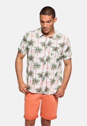 Hombre con camisa de manga corta con estampado de palmeras verdes y pantalones cortos coral con dobladillo vuelto, de pie sobre fondo blanco.