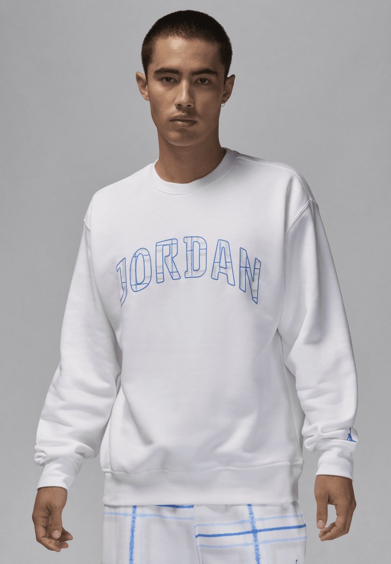 Sweatshirt blanc avec "JORDAN" en lettres superposées bleues, col rond côtelé et manches longues. Détail logo subtil sur le poignet gauche.