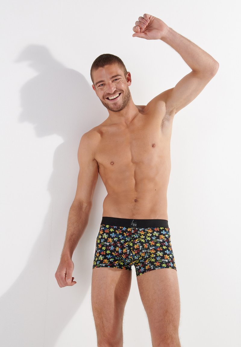 Black boxer briefs featuring a multicolored floral pattern, with a contrasting black waistband. Model poses with an arm raised, smiling.