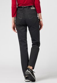 Pantaloni in denim neri con un design aderente, caratterizzati da cuciture decorative sulle tasche posteriori e una cintura nera lucida. Indossati con scarpe nere e dorate.