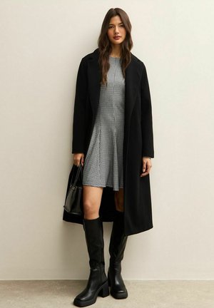 Cappotto lungo nero su un vestito a quadri, stivali neri al ginocchio con tacco quadrato e una piccola borsa nera. Texture lisce e forme definite.