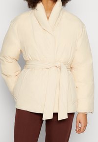 Vadderad gräddfärgad jacka med sjalkrage, midjebälte och sidofickor, gjord av mjukt tyg med en slät textur.