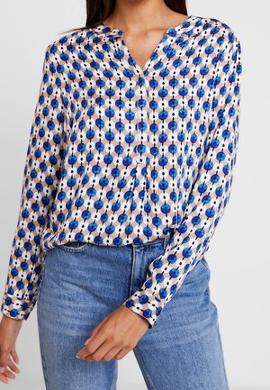 Kobieta ubrana w bluzkę z długim rękawem w niebiesko-czarny okrągły kwiatowy wzór, połączoną z dopasowanymi do talii niebieskimi dżinsami, z rozluźnionymi rękami.