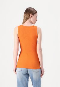 Top de tirantes naranja acanalado con escote redondeado, que muestra un ajuste slim. Llevado con jeans de corte relajado en un tono azul claro, con un tatuaje en el brazo.