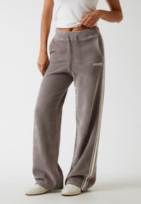 Szare szerokie dresy wykonane z miękkiego materiału, z białymi paskami po bokach oraz logo "GUESS" na lewej biodrze. Pas ze sznurkiem.