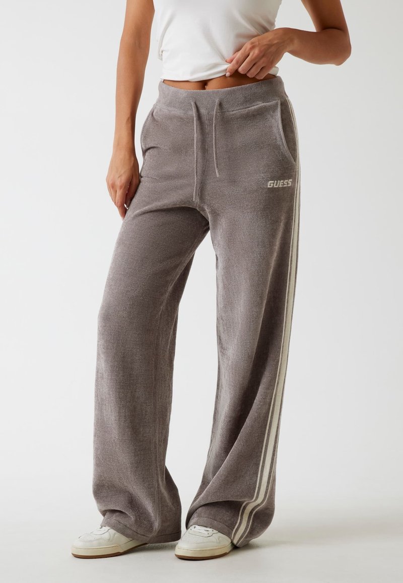 Szare szerokie dresy wykonane z miękkiego materiału, z białymi paskami po bokach oraz logo "GUESS" na lewej biodrze. Pas ze sznurkiem.