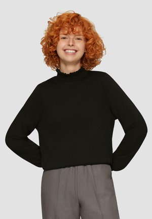 Zwarte trui met een hoge gerimpelde kraag, lange mouwen en een relaxte pasvorm, gedragen met grijze broek, gepresenteerd tegen een effen achtergrond.
