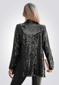 Zwarte pailletten blazer met een gestructureerd oppervlak, lange mouwen en een op maat gemaakte kraag; gecombineerd met een lichtgekleurde clutch.