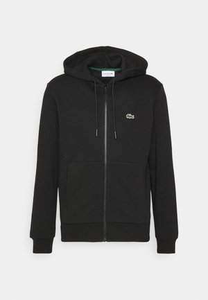 Črna zip-up kapuca iz mehke tkanine, s sprednjim žepom, rebrastimi manšeti in majhnim zelenim Lacoste logotipom na prsih.