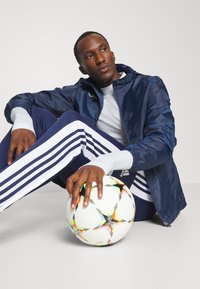 Navy windbreaker with elastic cuffs, paired with navy track pants featuring white stripes. A white soccer ball with colorful accents is held.
