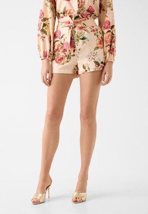 Femme portant un haut à manches longues beige à motif floral et un short taille haute assorti avec une ceinture nouée, associé à des mules dorées à talons hauts.