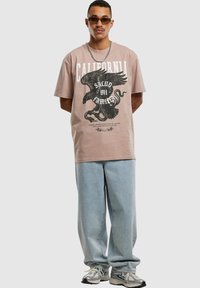 Lyserød t-shirt med korte ærmer og sort ørne grafik samt teksten "CALIFORNIA", parret med løse lyseblå denim jeans og grå sneakers.