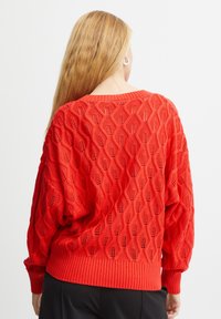 Pulover tricotat roșu cu un model texturat în diamant, având un tiv și manșete cu nervuri. Designul este oversized, cu un decolteu rotund.