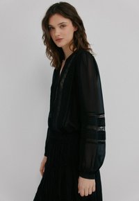 Blouse noire transparente à manches longues, avec des accents en dentelle et un col en V profond. Présente des poignets froncés et des motifs à rayures complexes sur les manches.