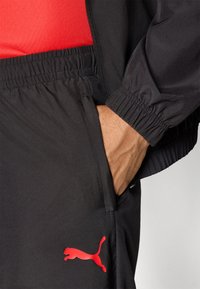 Črne športne hlače z elastiko v pasu, ki imajo eleganten rdeč Puma logotip na strani ter stranski žep za priročnost.