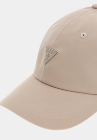 Gorra de béisbol beige hecha de tela suave, con un detalle en forma de triángulo de logo plateado y costuras visibles en el borde.