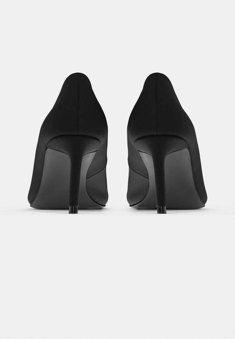 Une paire de chaussures à talons hauts noirs montrée de l'arrière sur une surface et un fond blancs.