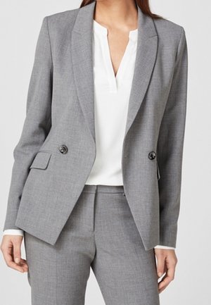 Kvinde iført en skræddersyet grå blazer med sorte knapper over en hvid V-hals bluse og matchende grå bukser.