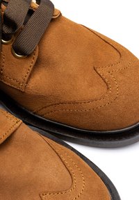 Chaussures en daim marron avec une texture lisse, coutures jaunes contrastantes, semelle ronde noire et coutures décoratives sur l\'orteil.