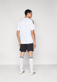 Bijeli sportski dres s crnim prugama na ramenima, uparen s crnim kratkim hlačama i bijelim čarapama do koljena s tri crne pruge.