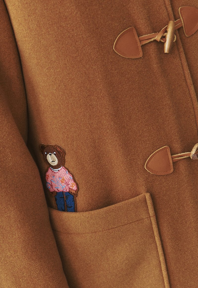 Brązowy wełniany płaszcz z zapięciem na togglowe zapięcia, z haftem niedźwiedzia na różowym swetrze i niebieskich dżinsach na lewej kieszeni.