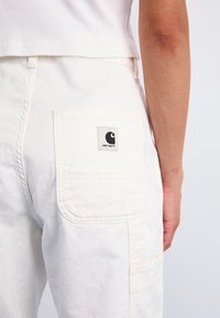 Hvite cargo-bukser laget av tykk bomull, med en baklomme med logo-patch og synlige sømdetaljer. Myk, strukturert stoff.