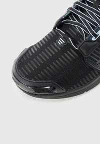 Fekete sportcipő közeli kép, amelyen texturált hálós felsőrész, fényvisszaverő fűzők és megerősített orr-rész látható, egyszerű világos háttér előtt.