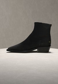 Black ankle boot with pointed toe and low angled heel, displayed on a round beige surface against a neutral gray background.