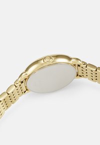 Guldmetall damklocka med rund boett, slät guldtonad bezel och en vävd armbandsrem med spänne.