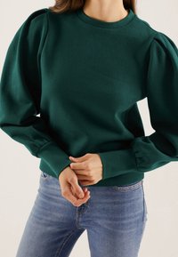 Groene trui met pofmouwen, ronde hals en geribbelde boorden en zoom. Draagt een blauwe spijkerbroek met een gladde stofstructuur.