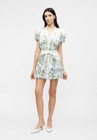 Woman wearing a short, white floral dress with puff sleeves, tied waist belt, and white pointed heels, standing against a plain background.