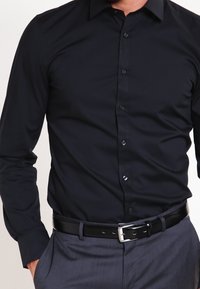 Zwarte langemouwen shirt met een aansluitend ontwerp, voorzien van een knoopsluiting aan de voorkant en een subtiele glans, gecombineerd met een zwarte riem en grijze broek.