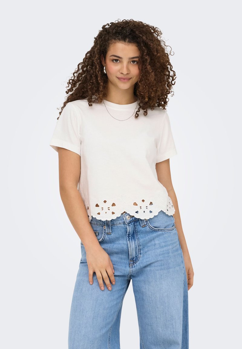 Jeune femme aux cheveux bouclés portant un top blanc à manches courtes avec un ourlet en dentelle festonné et un jean taille haute bleu clair.