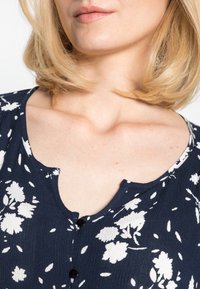 Blusa floral azul-marinho com padrões de flores brancas, decote redondo, detalhe de botão e tecido texturizado. Apresenta mangas curtas e caimento suave.