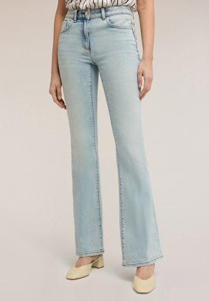 Femme portant un jean flare taille haute bleu clair, une chemise rayée rentrée et des chaussures beiges à talons blocs, debout devant un fond uni.
