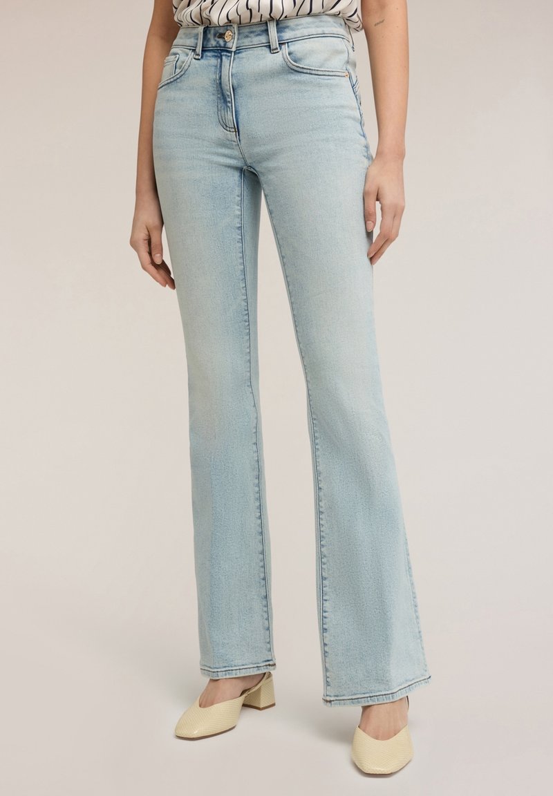 Femme portant un jean flare taille haute bleu clair, une chemise rayée rentrée et des chaussures beiges à talons blocs, debout devant un fond uni.