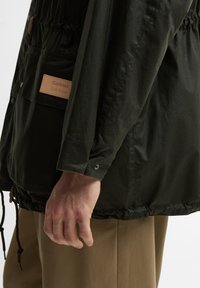 Άνδρας που φοράει σκούρο πράσινο μπουφάν Barbour με δερμάτινο ταμπελάκι λογότυπου, εμφανιζόμενος από το πλάι με έμφαση στο μανίκι, την τσέπη και το χέρι· εμφανίζονται μπέζ παντελόνια.