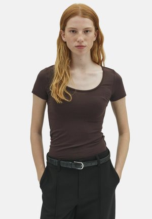 Jeune femme aux longs cheveux roux et aux taches de rousseur porte un haut marron ajusté à manches courtes et un pantalon noir avec une ceinture noire, les mains dans les poches.
