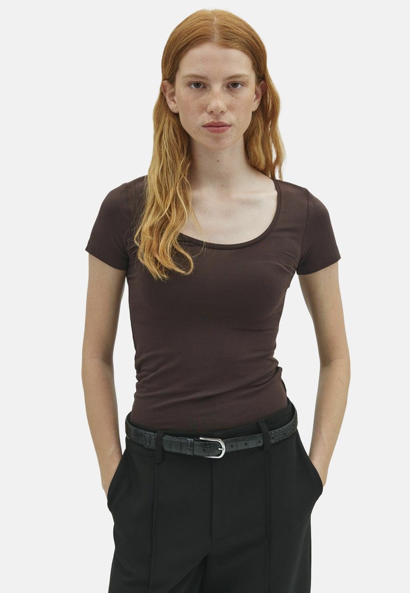 Jeune femme aux longs cheveux roux et aux taches de rousseur porte un haut marron ajusté à manches courtes et un pantalon noir avec une ceinture noire, les mains dans les poches.