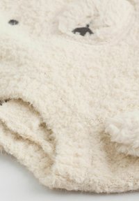 Tkanina w kolorze kremowym, o miękkiej fakturze, z futrzanymi akcentami przypominającymi twarz i uszy niedźwiedzia; posiada wyszywane oczy i zaokrąglony kształt.
