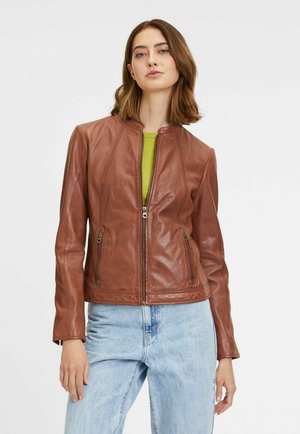 Giacca in pelle marrone con design aderente, colletto alla coreana e chiusura frontale con zip. Presenta tasche con zip e finitura texturizzata. Indossata sopra una maglia verde.