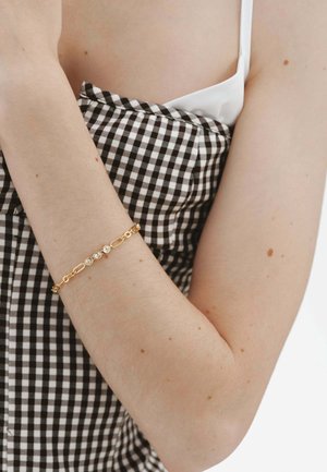 Brazo con ropa de cuadros en blanco y negro y una pulsera dorada con cuatro pequeñas piedras claras en la muñeca.