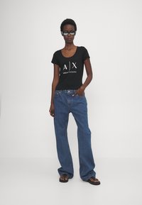 Čierne priliehavé tričko s bielym logom "A|X ARMANI EXCHANGE" v kombinácii s vysokými modrými džínsami so širokými nohavicami a čiernymi žabkami.