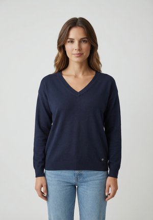 Donna con capelli castano ondulati che indossa un maglione blu navy a scollo a V e jeans azzurri, in piedi davanti a uno sfondo chiaro uniforme.